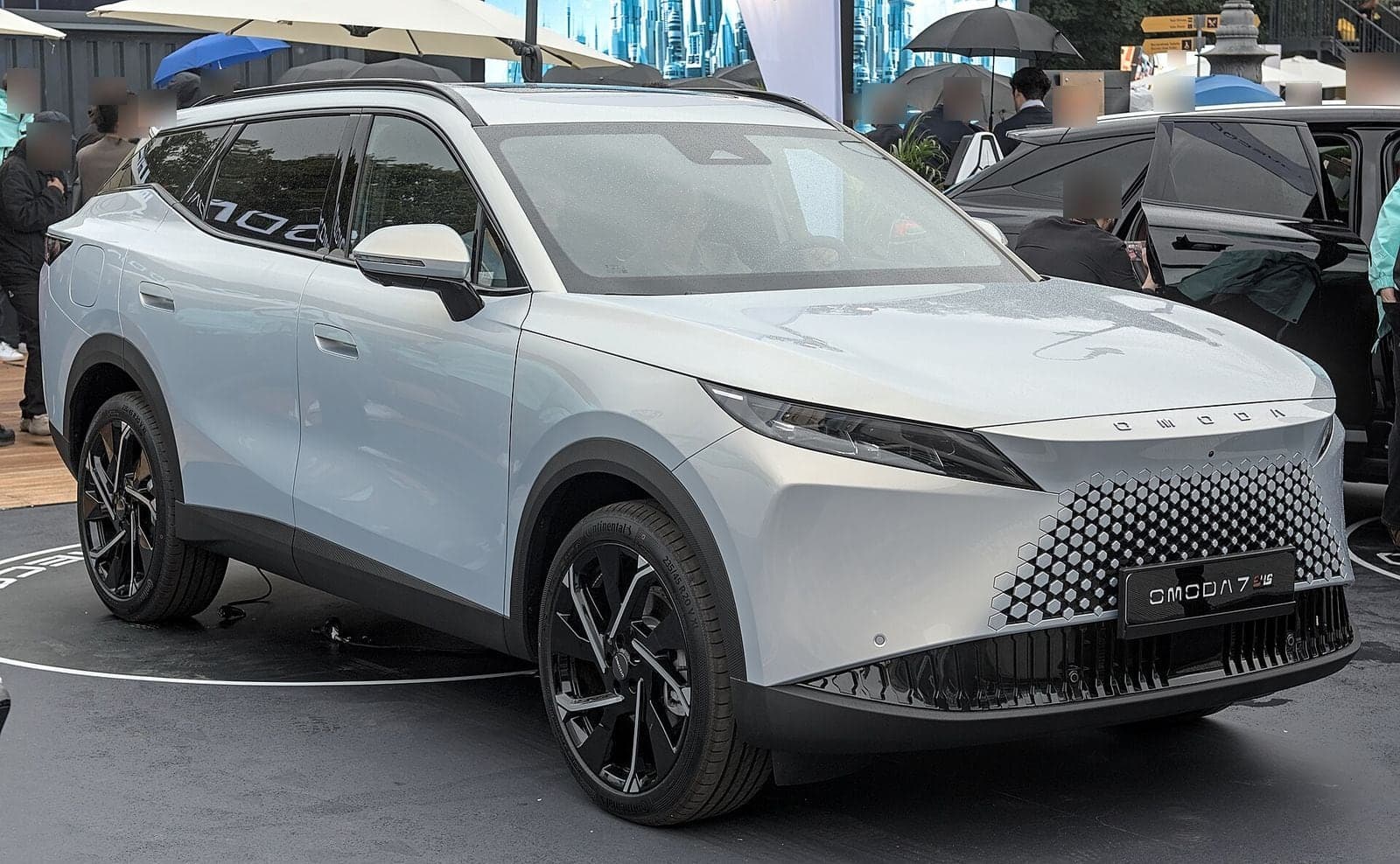 Hellgraues SUV Omoda 7 SHS mit Kühlergrill aus sechseckigen Elementen auf einem Messestand im Freien, im Hintergrund Besucher mit Schirmen
